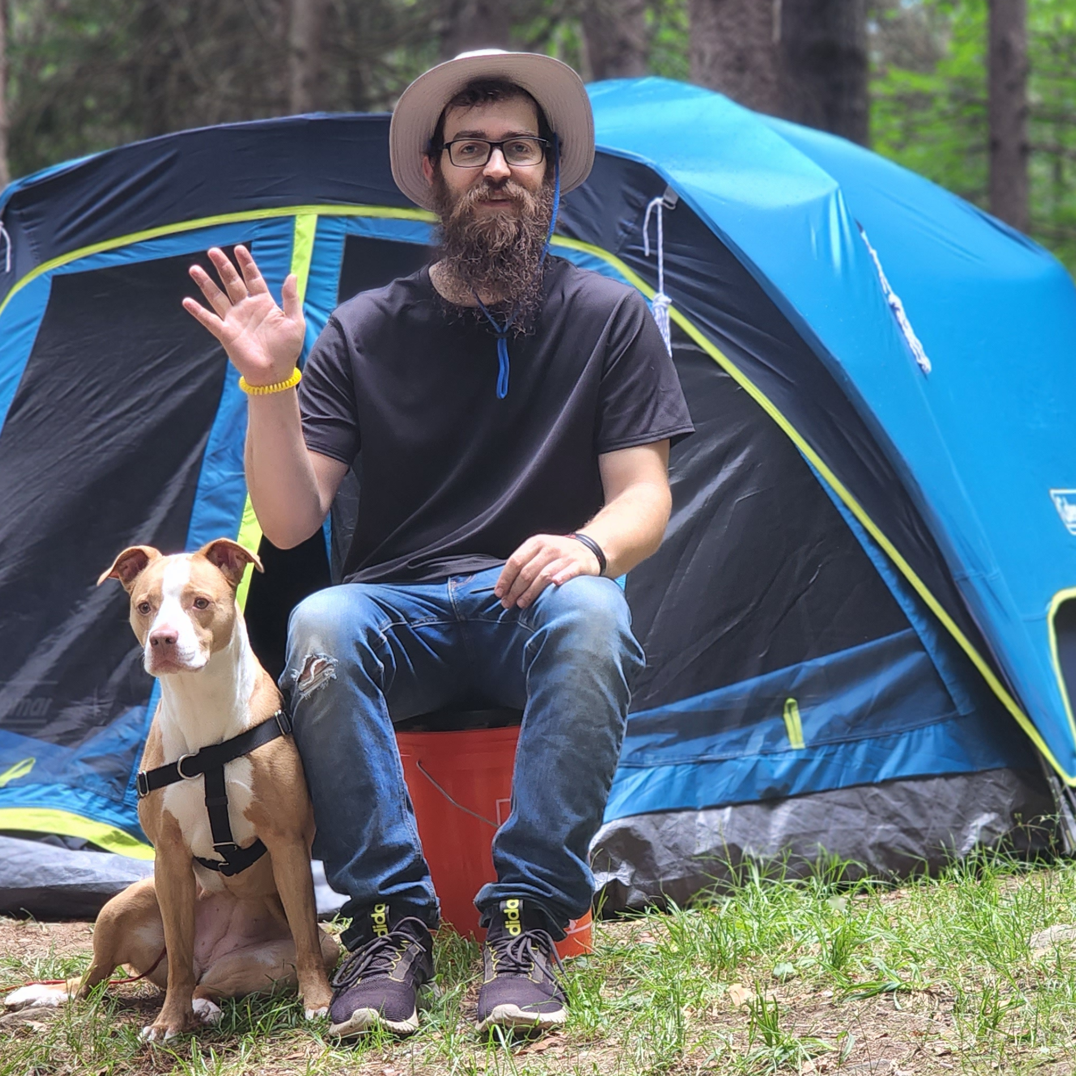 The image shows a bearded man wearing a hat sitting in front of a blue tent. The man is waving and holding the leash of his light brown and white pitbull boxer mix dog, also pictured.